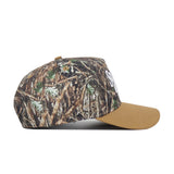 The Scheels Camo Explorer Hat is shown from the side on a white background, featuring a brown camo-patterned crown with grass and leaves, solid tan brim, structured fit, and high-quality cotton construction.