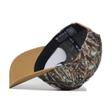 The Scheels Camo Explorer Hat is shown on its side, highlighting the inside label and black sweatband. It features a brown brim and a crown with a green, brown, and tan camo pattern.