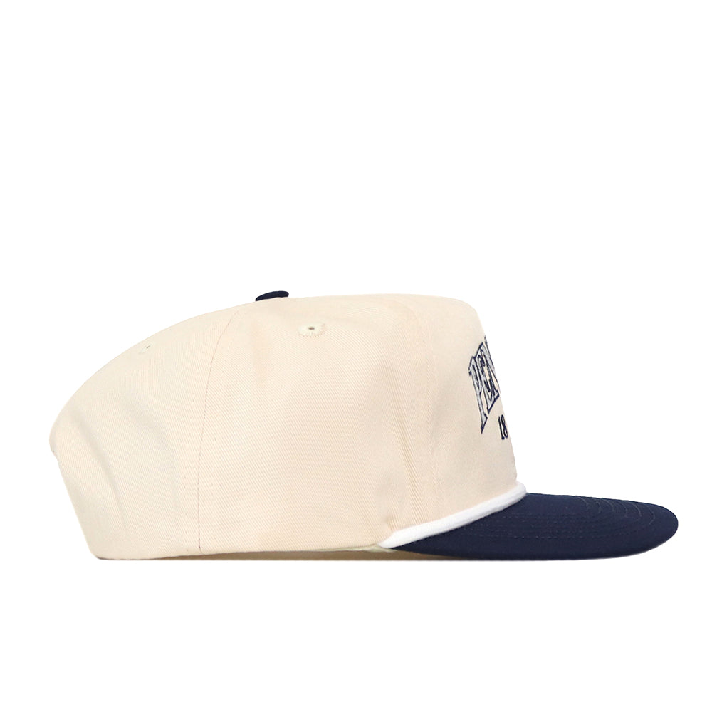 A side view of the Penn State Vintage Block Hat by No Rivals showcases its cream-colored snapback design with a navy blue brim. The hat features an embroidered logo, a small button on top, and partially visible text on the front panel. This officially licensed snapback is ideal for fans who value both quality and style.