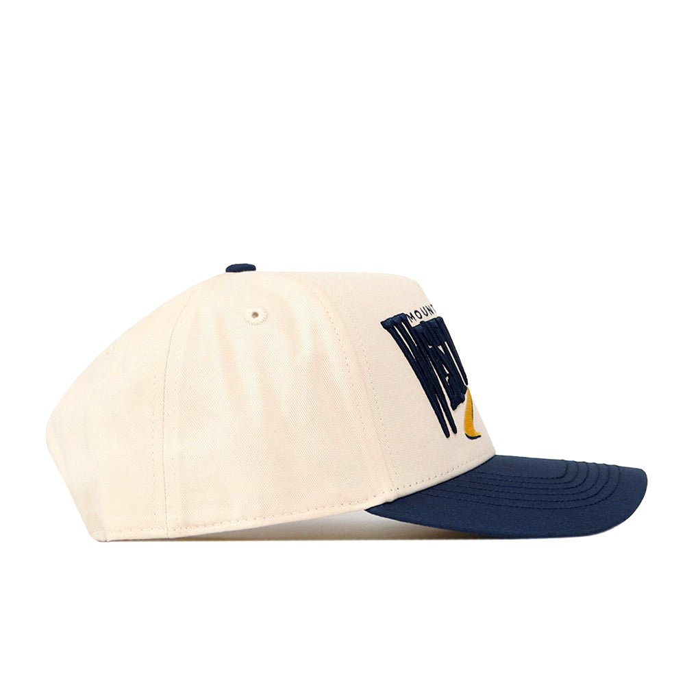 The No Rivals WVU Mountaineers Stride Hat is a cream-colored baseball cap with a navy blue brim, shown from the side. This structured fit cap is crafted from high-quality cotton and features a snapback closure. The front panel displays a navy and yellow embroidered design.