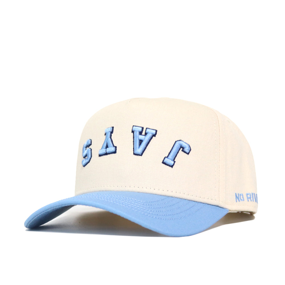 The Upside Down JAYS Hat by No Rivals features a cream body, light blue brim, and structured fit. "JAVS" is embroidered upside down in large light blue letters on the front, with "NO RIV" stitched in smaller blue text on the brim’s side.