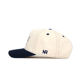 Side view of the cream-colored No Rivals ETSU Vintage Hat with a black brim, crafted from premium cotton. Features black stitched eyelets, an embroidered NR logo on the side, and an adjustable snapback closure.