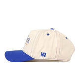 The Air Force Vintage Hat by No Rivals is displayed in a side view, showcasing its beige and blue design. Made from high-quality cotton, this structured cap features blue stitching, a blue brim, and “NR” embroidered near the back.