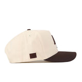 Introducing the Air Force Espresso Hat by No Rivals: a cream-colored snapback with a contrasting dark brown brim, displayed from a side angle. This cap boasts a small brown square patch on the side panel, ventilation eyelets for breathability, and offers a structured fit for comfort and style.
