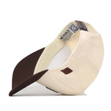 The Air Force Espresso Hat by No Rivals is pictured upside down, displaying its cream-colored crown, brown brim, and high-quality cotton construction. The inside of the cap reveals tags with text and a QR code.