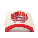 The No Rivals Nebraska Cornhuskers Stadium Hat is a white and red snapback featuring “Nebraska Cornhuskers” embroidered on the front above an image of a football stadium, with officially licensed logos for a sporty, authentic style.
