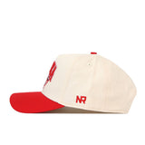 The No Rivals Nebraska Cornhuskers Stadium Hat is a cream-colored, officially licensed snapback baseball cap featuring a red brim and an embroidered red NR logo on the side. Displayed from a side view against a white background, the detailed embroidered logos stand out on this stylish fan accessory.