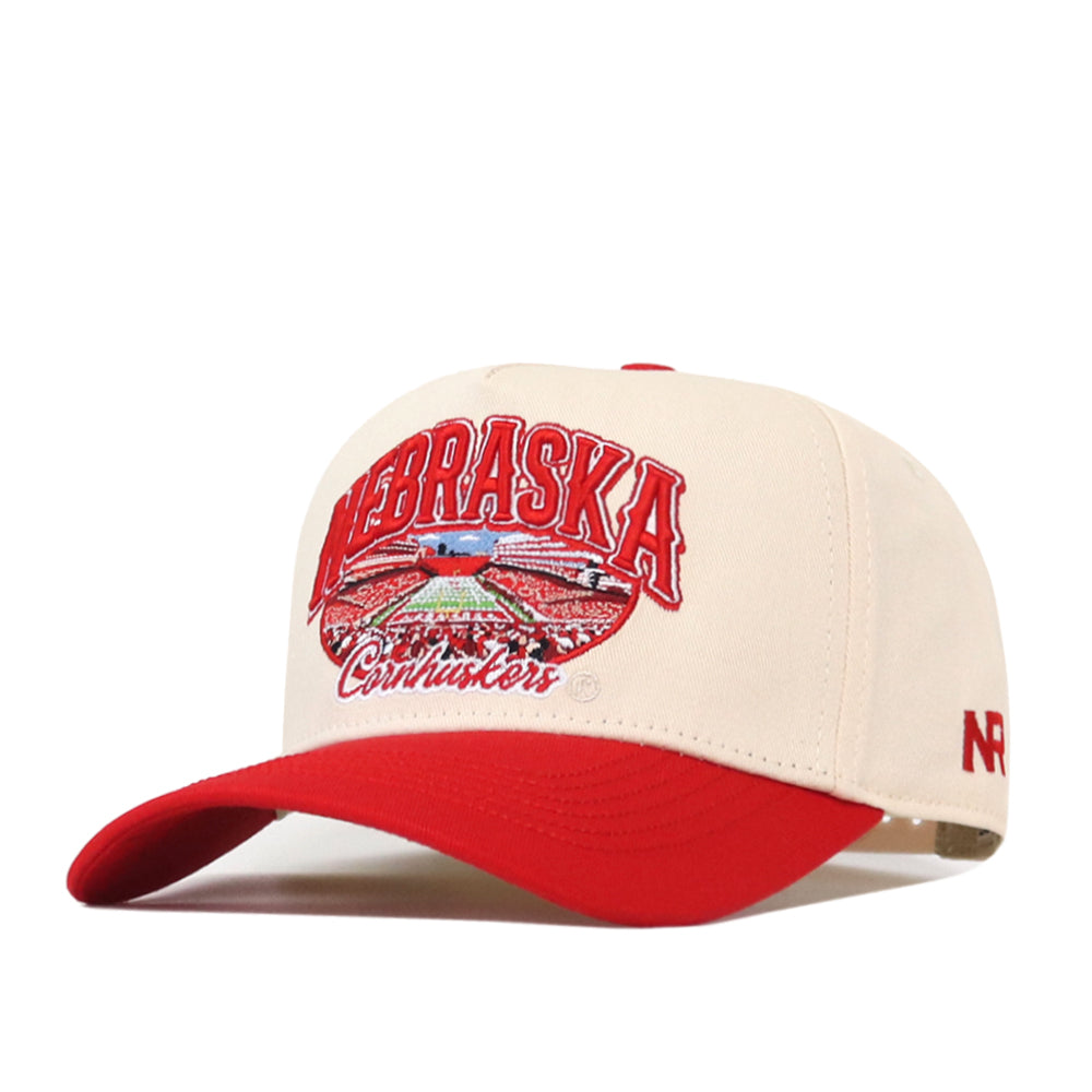 The No Rivals Nebraska Cornhuskers Stadium Hat is a beige and red snapback featuring embroidered Nebraska Cornhuskers text and a football stadium on the front, officially licensed logos, a vibrant red brim, and a bold red “NR” on the side.