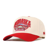 The No Rivals Nebraska Cornhuskers Stadium Hat is a beige and red snapback featuring embroidered Nebraska Cornhuskers text and a football stadium on the front, officially licensed logos, a vibrant red brim, and a bold red “NR” on the side.