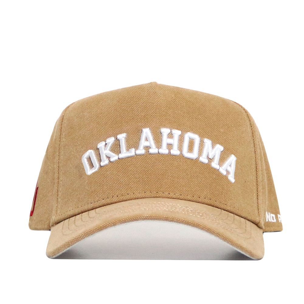 The Oklahoma Rustic Hat by No Rivals is a tan cotton canvas hat with a structured fit, featuring "OKLAHOMA" embroidered in white arched letters on the front.