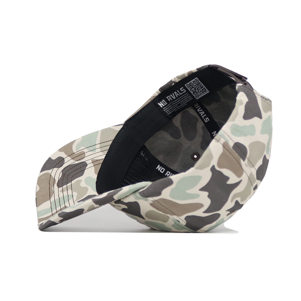 The Montana Huntsman Hat by No Rivals lies on its side, showing the black snapback strap and NO RIVALS label inside. This 5-panel hat features classic duck camo in shades of green, brown, and beige.