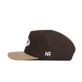 The Texas A&M Neutral Patch Hat by No Rivals is a brown snapback with a beige brim, a front embroidered white oval patch, and "NR" in white on the side for a structured fit.
