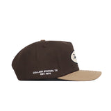 The Texas A&M Neutral Patch Hat by No Rivals is a brown snapback with a tan brim, featuring white "COLLEGE STATION, TX EST. 1876" embroidery on the side and a white oval patch on front. This officially licensed cap has a structured fit.