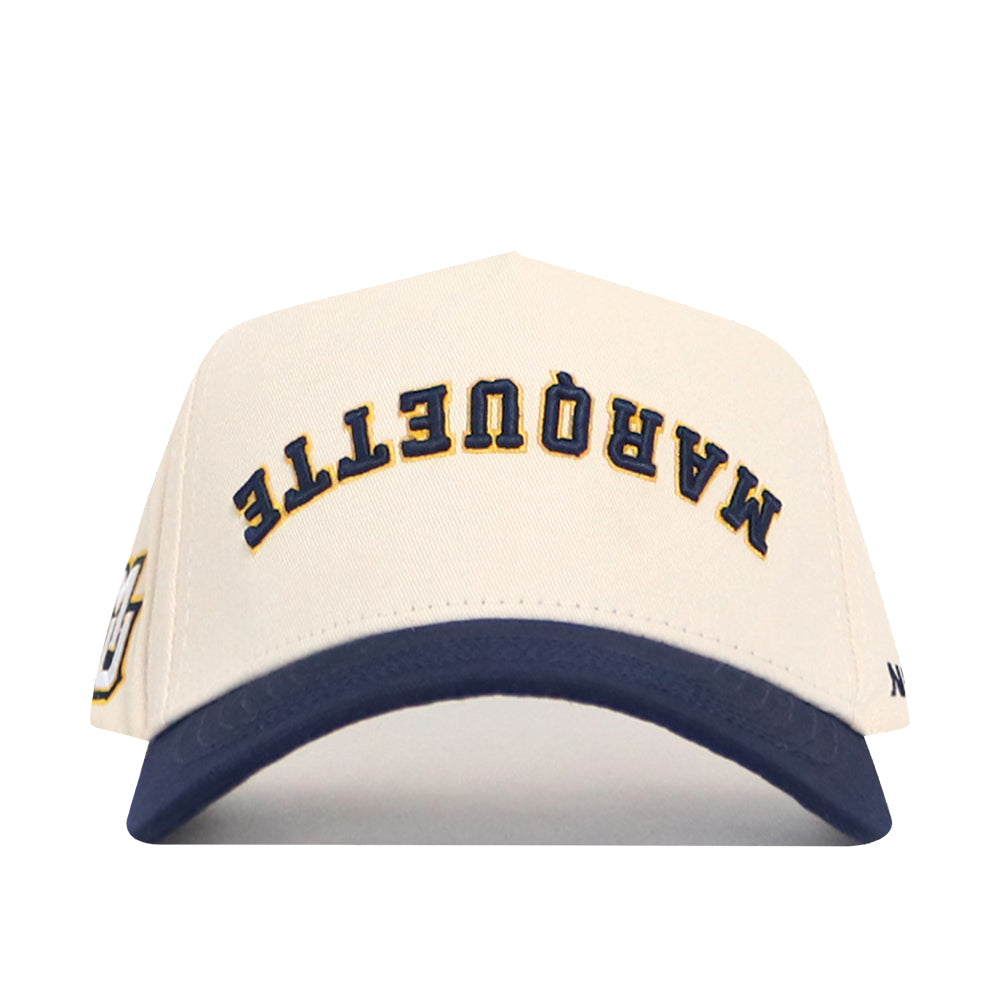 The Upside Down MARQUETTE Hat by No Rivals is a white cotton-canvas snapback with a navy blue brim, featuring "MARQUETTE" embroidered in yellow and navy blue across the front.