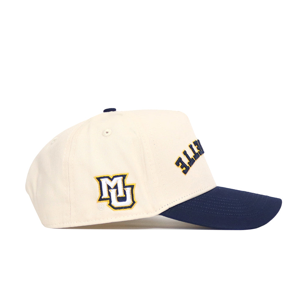 The Upside Down MARQUETTE Hat by No Rivals is a cream cotton-canvas baseball cap with a navy brim, an embroidered MU logo on the side, partial text on the front, and structured for all-day comfort.