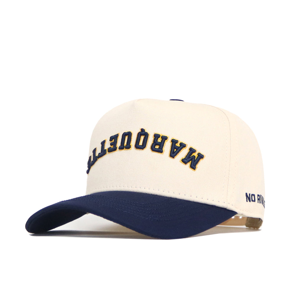 The Upside Down MARQUETTE Hat - Classic Colors by No Rivals is a cream cotton-canvas snapback with a navy brim, featuring “MARQUETTE” embroidered upside down in navy and yellow on the front, and "NO" on the left side of the brim.