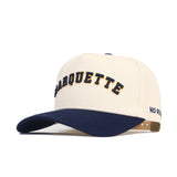 The No Rivals Right Side Up MARQUETTE Hat - Classic Colors is a cream cotton-canvas snapback with "MARQUETTE" in navy and gold on the front, a navy bill, and "NO REST" stitched on the left side.