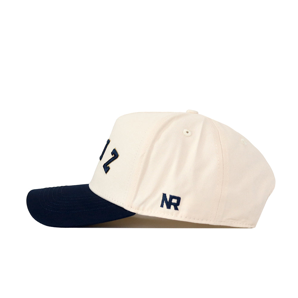A cream-colored Akron Zips | Upside Down Zips Hat - Classic by No Rivals, featuring a navy blue brim, “NR” embroidery on the side, partially displayed front text, and a classic snapback design.