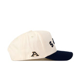 A cream-colored Akron Zips | Upside Down Zips Hat - Classic by No Rivals is shown from the side, featuring a navy blue brim. The side showcases an embroidered letter A in black and gold, evoking its Akron Zips inspiration. Partial black lettering appears on the front of this stylish snapback.