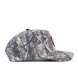 Side view of the Auburn Outland Hat by No Rivals, a baseball cap with a camouflage pattern in earthy tones and tree branch designs. The bill is slightly curved, and the front displays a partially visible small red and black embroidered logo. This snapback combines rugged outdoor style with classic comfort.