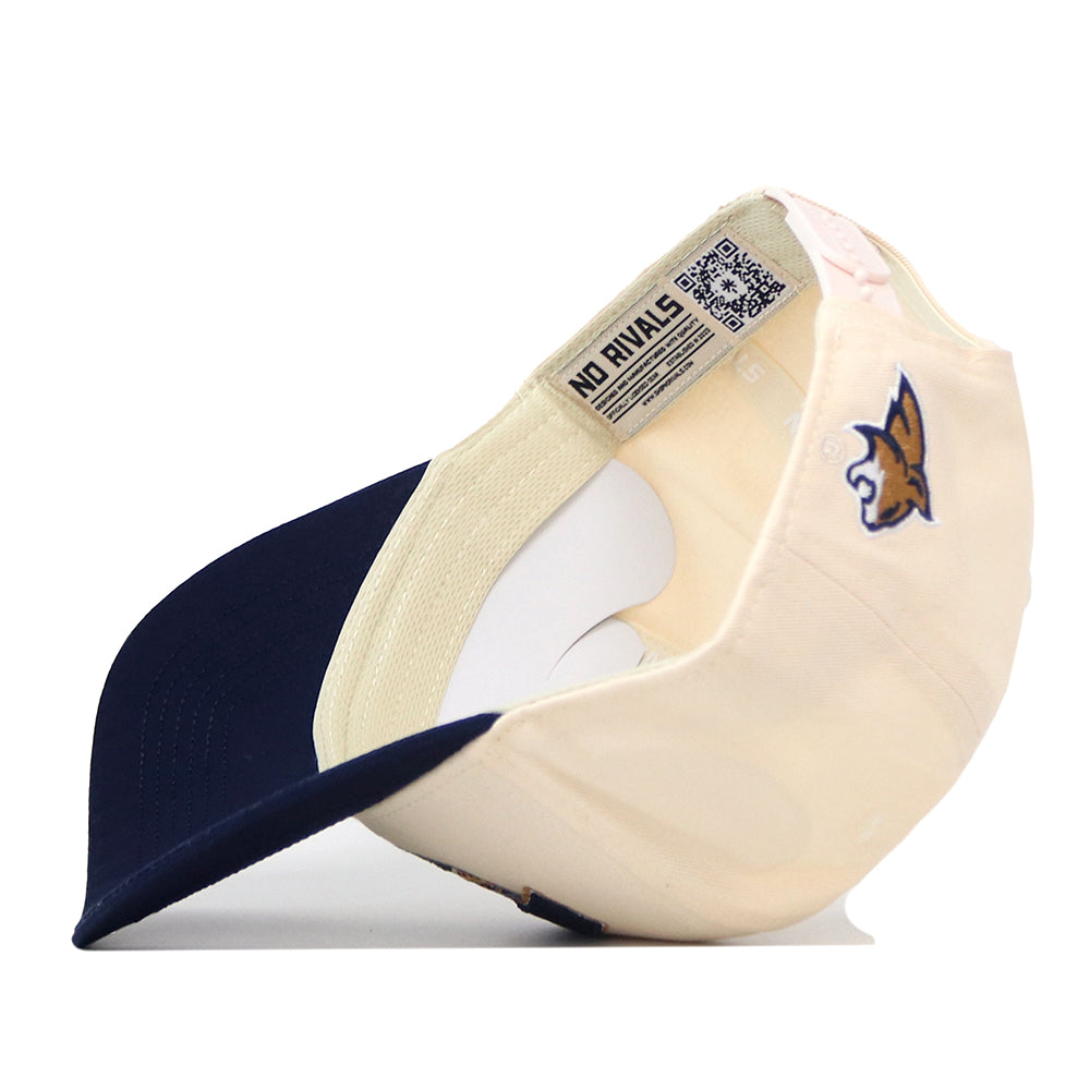 A cream and navy blue Montana State Bobcats Stadium Hat by No Rivals is flipped upside down, displaying the inner tags, a QR code, “NO RIVALS” text, and officially licensed embroidered logos—including a wildcat logo on the side.