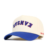 The Upside Down KANSAS Hat by No Rivals features a cream crown, blue brim, “KANSAS” upside down in blue with red outline on the front, “NO RIV” stitched in red on the side, and is made from high-quality cotton for comfort and durability.