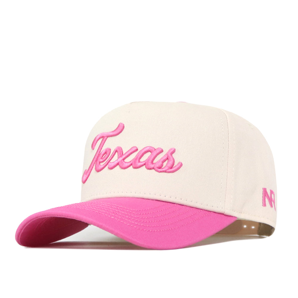 The Texas Pink Out Hat by No Rivals is a cream-colored baseball cap featuring “Texas” in pink script on the front and a pink brim. This officially licensed snapback breast cancer hat includes small pink initials on the side.