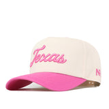The Texas Pink Out Hat by No Rivals is a cream-colored baseball cap featuring “Texas” in pink script on the front and a pink brim. This officially licensed snapback breast cancer hat includes small pink initials on the side.