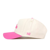 The Dallas Pink Out Hat by No Rivals is a cream-colored baseball cap featuring a pink brim and pink embroidered “NR” lettering on the side. It includes a convenient snap back adjustment and is displayed in a side profile view against a white background.