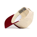 The Alabama Stride Hat by No Rivals is a beige and red cotton baseball cap with a structured fit, curved dark red bill, and an inner label reading NO RIVALS plus a QR code on the band. The photo shows it flipped to display these details.
