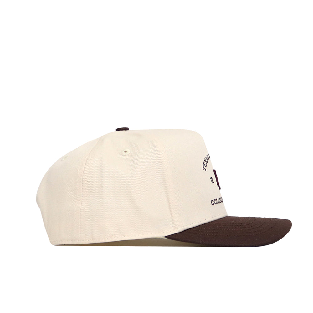 The No Rivals Texas A&M Outlaw Hat - Classic is a cream-colored snapback with a brown brim, shown in side profile. Made from quality cotton, it features partial front-left text and a small logo.