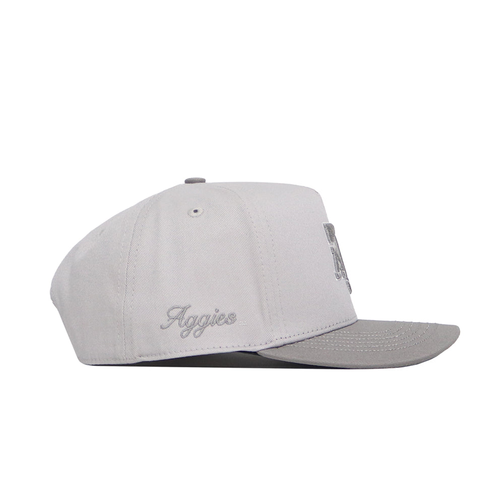 The No Rivals Texas A&M Graphite Hat is a structured, light gray snapback with a slightly darker flat brim. Made from premium cotton, it features "Aggies" script embroidery on the left side and is shown in profile, facing right.