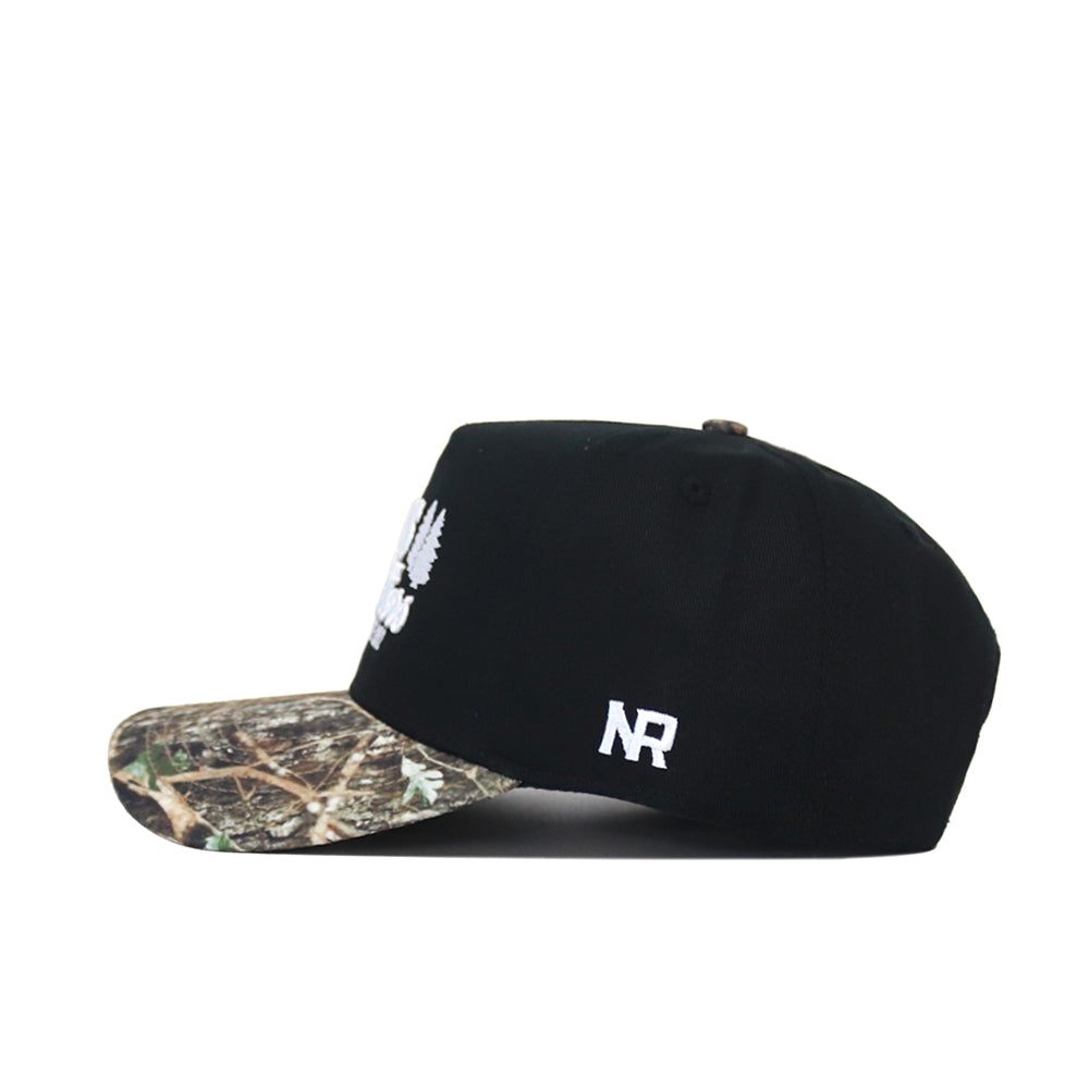 The Scheels Camo Trailblazer Hat is a black snapback with a camo brim, white embroidered front and side logos, made from high-quality cotton for a structured fit. Shown in side profile.