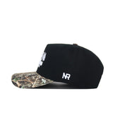The Scheels Camo Trailblazer Hat is a black snapback with a camo brim, white embroidered front and side logos, made from high-quality cotton for a structured fit. Shown in side profile.