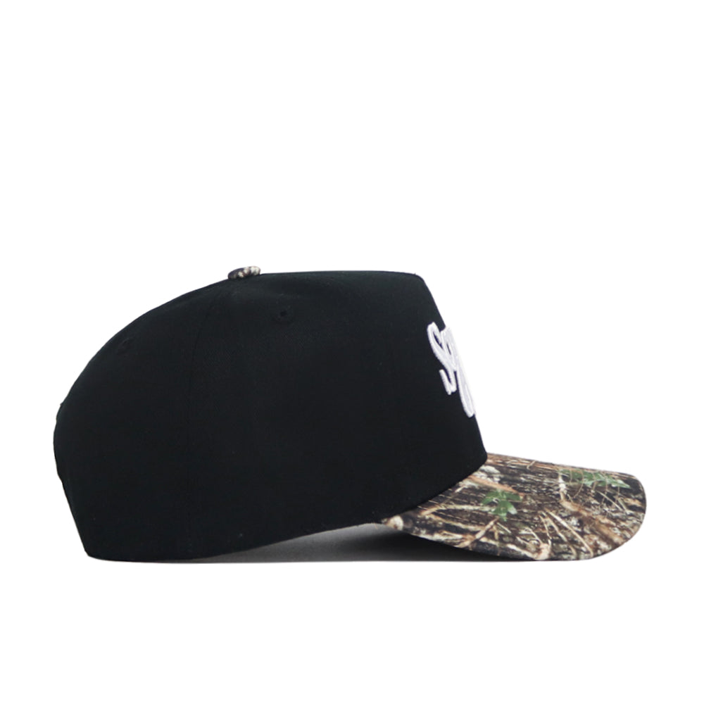 The Scheels Camo Trailblazer Hat is a black snapback with a camo-patterned brim, shown side-on against a white background. Made of high-quality cotton, it features partially visible white embroidered lettering on the front panel.