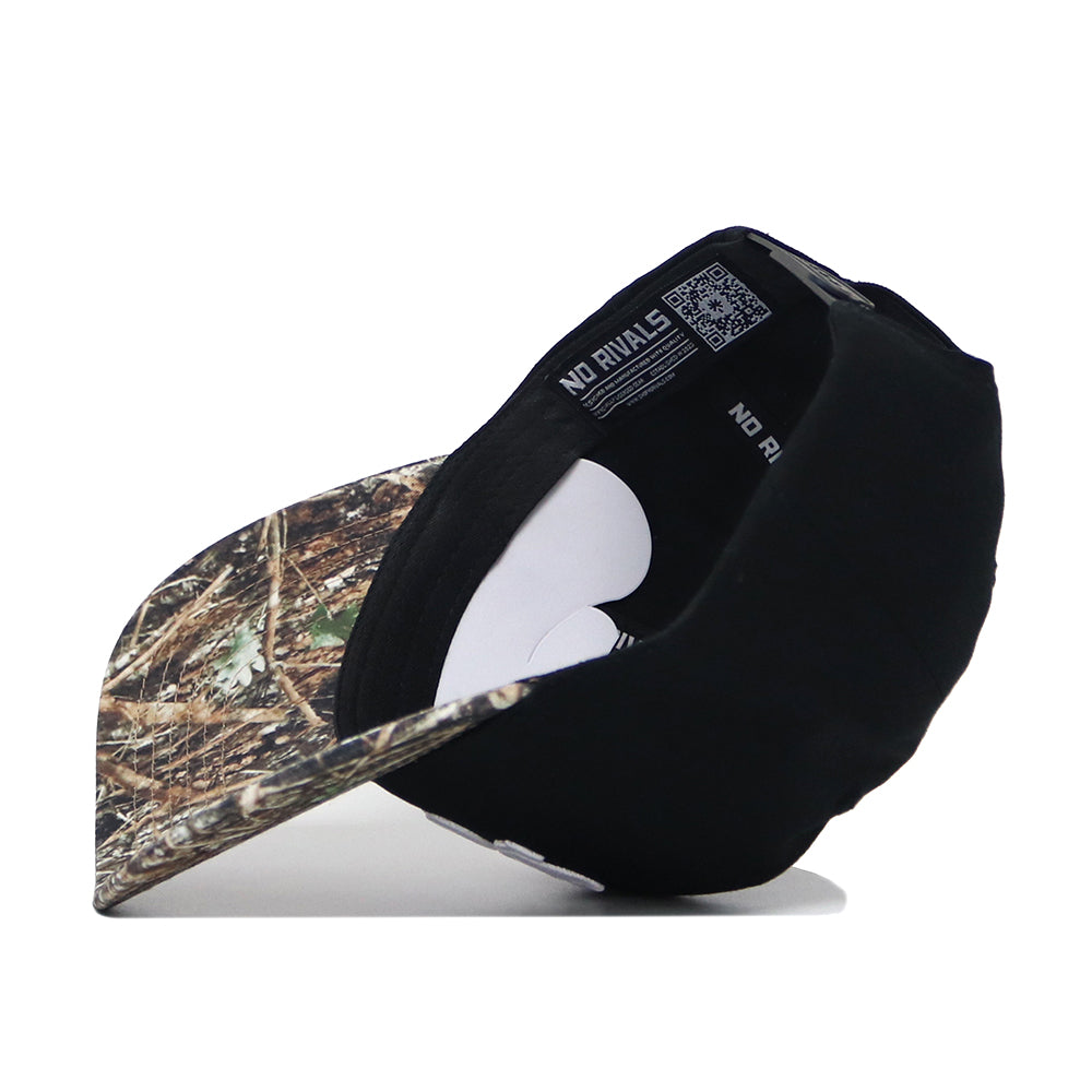 The Scheels Camo Trailblazer Hat is shown upside down, revealing "NO RIVALS" text and a QR code on the inner band. Its structured brim features a high-quality forest camouflage pattern in black and camo cotton.