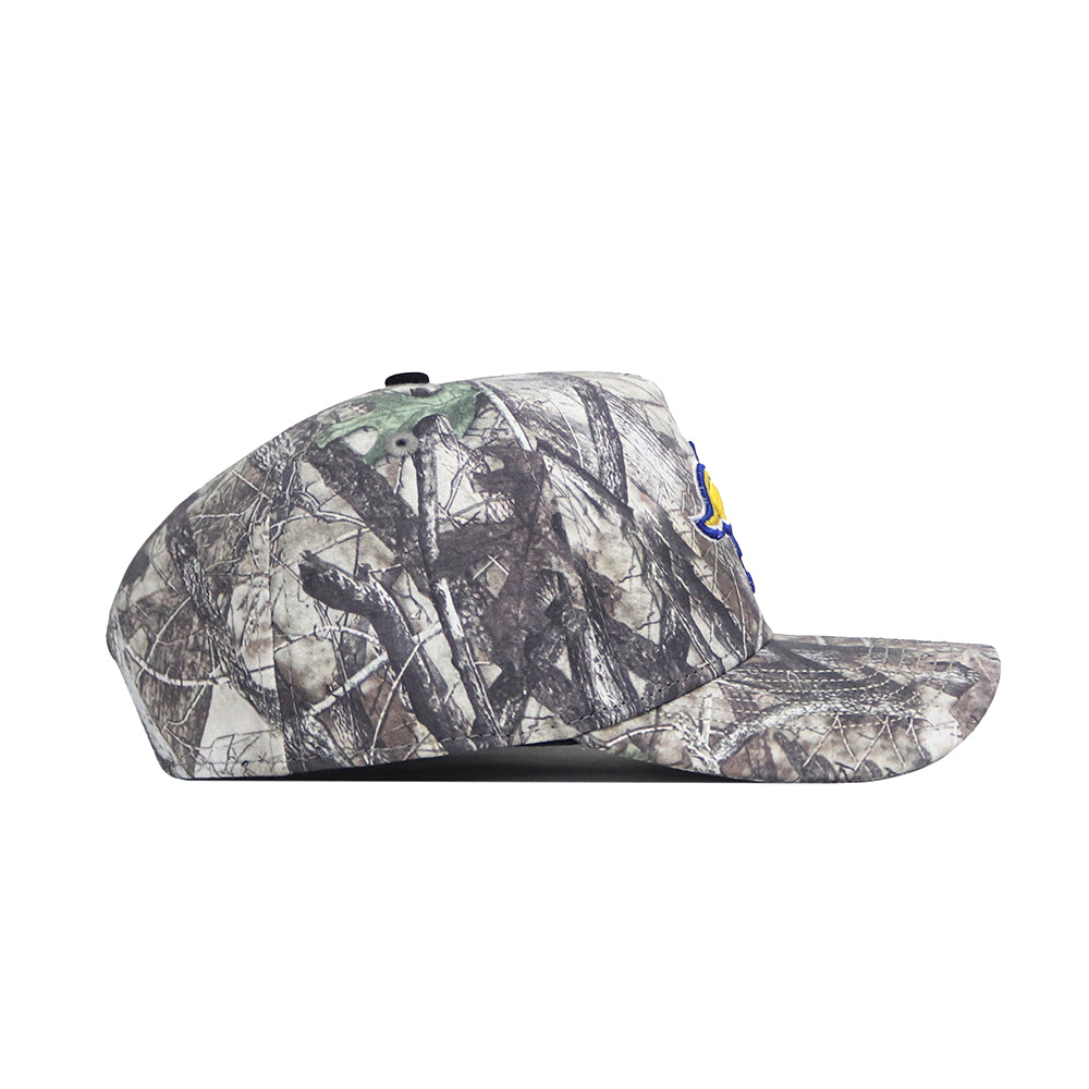 A side view of the No Rivals South Dakota State Outland Hat, a flat-brim snapback with a forest camo pattern featuring tree branches and leaves in brown, green, and gray. Made from high-quality cotton with a structured fit.