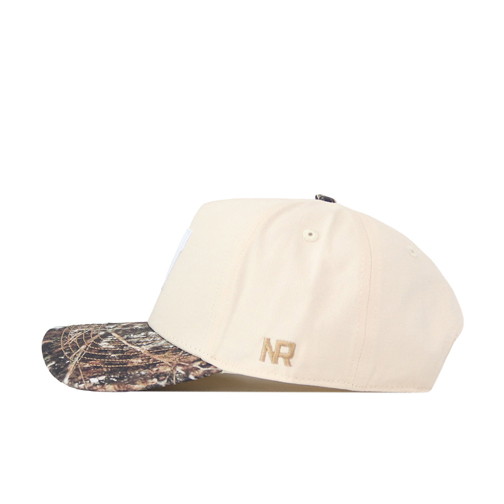 A beige Wisconsin Camo Connector Hat by No Rivals, featuring a camouflage-patterned brim and side view. The gold-embroidered NR initials on the lower left crown add a signature touch to your gameday fit.