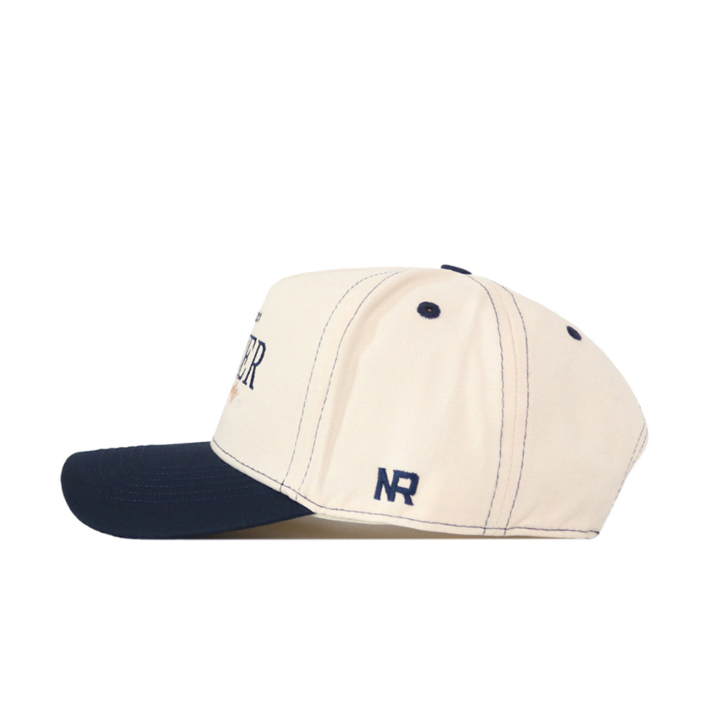 A cream-colored Xavier Musketeers Vintage Hat by No Rivals, viewed from the side, featuring a dark navy brim, ventilation holes, and an embroidered NR logo in navy on the side—making it a perfect addition to your gameday fit.
