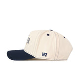 A cream-colored Xavier Musketeers Vintage Hat by No Rivals, viewed from the side, featuring a dark navy brim, ventilation holes, and an embroidered NR logo in navy on the side—making it a perfect addition to your gameday fit.