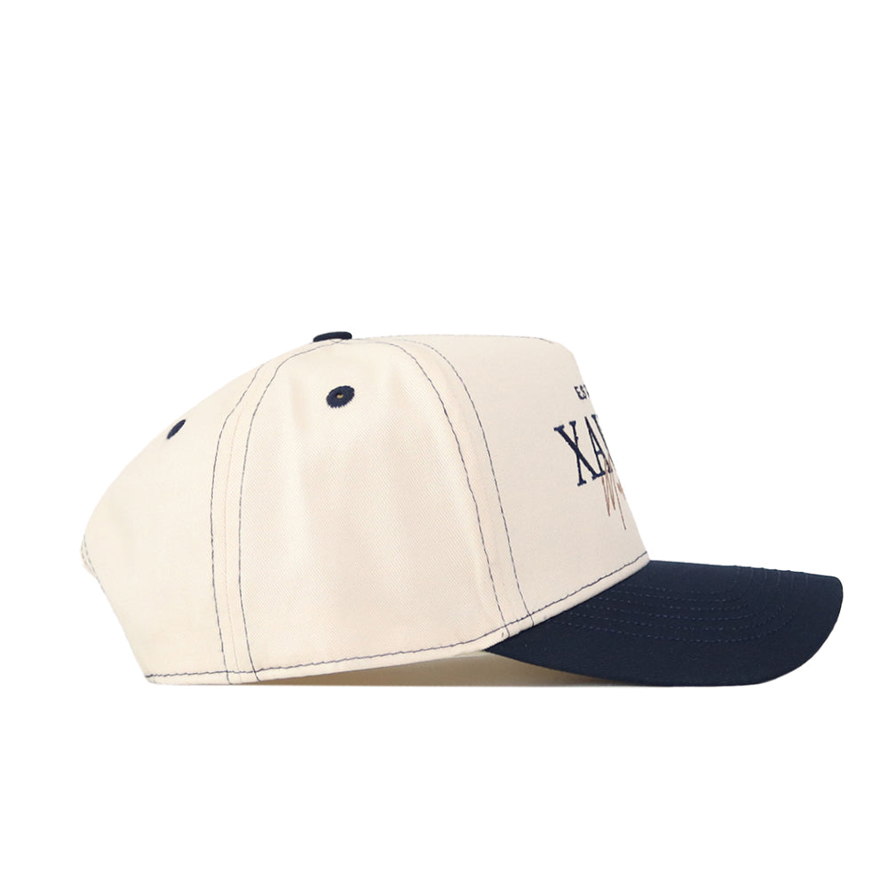 The No Rivals Xavier Musketeers Vintage Hat, featuring a beige crown, navy blue brim, embroidered Xavier logo, and stitched eyelets, is pictured from the side against a white background—a classic snapback that’s perfect for your next gameday fit.