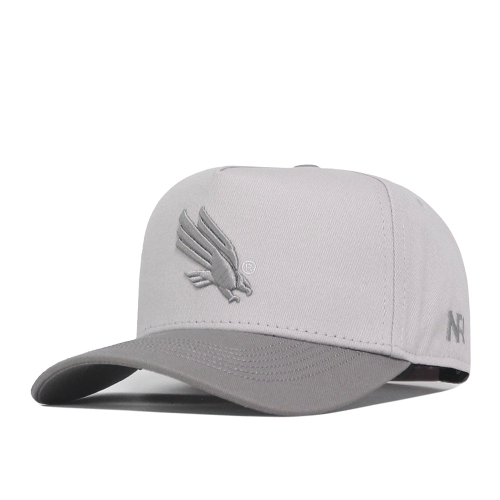 The No Rivals UNT Graphite Hat is a light gray baseball cap featuring an officially licensed embroidered bird logo on the front and a curved brim. It also has small stitched details, including letters, on the side of the cap.