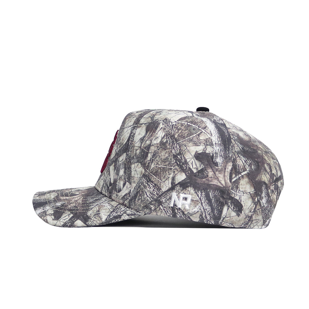 The Montana Outland Hat by No Rivals is a camouflage-print snapback with a flat brim, shown in side profile. It features a red embroidered front logo and white NP initials embroidered on the side.