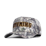 The No Rivals Wyoming Outland Hat is a structured cotton baseball cap with "WYOMING" boldly embroidered in yellow on the front. It features a curved brim and camouflage fabric in gray, brown, and green shades.