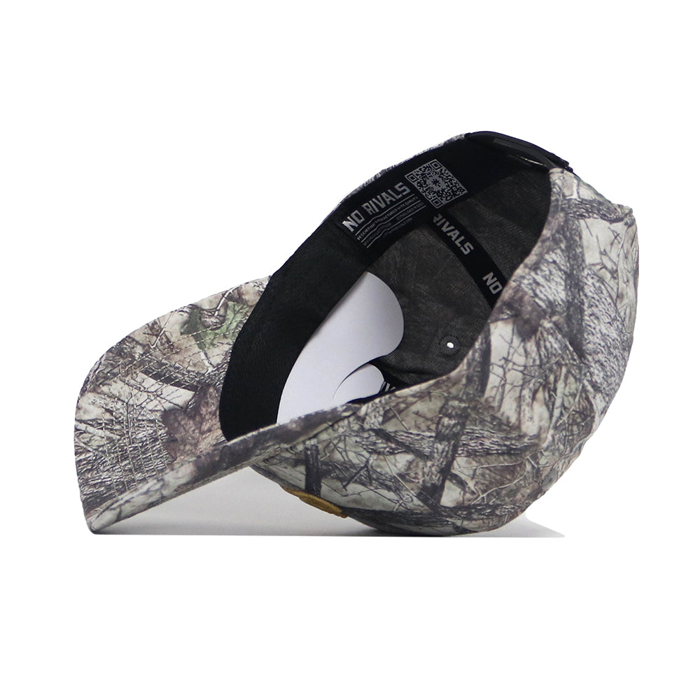 The JMU Outland Hat by No Rivals is a high quality cotton baseball cap in a woodland green and brown camouflage pattern, shown on its side to display the underside brim and interior band with "NO RIVALS" printed inside.
