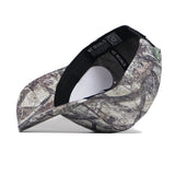 The Louisville Outland Hat by No Rivals is shown upside down, highlighting its black adjustable strap and interior tags. This high-quality cotton baseball cap features a green, brown, and tan forest camouflage pattern.