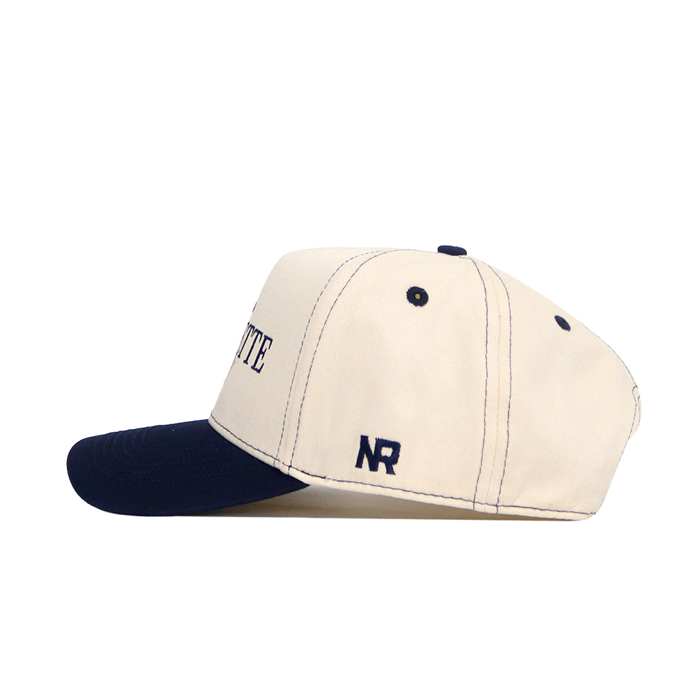 The No Rivals Marquette Vintage Hat features a cream snapback design with a navy brim, structured fit, navy stitching, and NR initials embroidered on the side. This high-quality cotton cap is displayed in side profile against a white background.