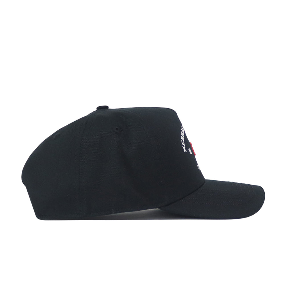 The Nebraska Outlaw Hat - Black by No Rivals is shown from the side, featuring a curved brim and visible stitching. Small embroidered logo text and a red square detail appear on the front panel, though the full text isn’t readable from this angle—perfect for adding a subtle touch to your gameday fit.
