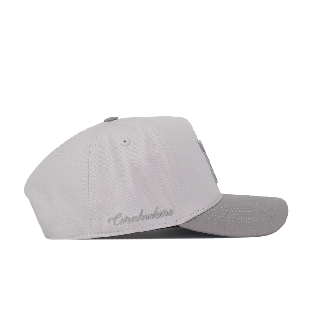 The No Rivals Nebraska Graphite Hat is a light gray baseball cap with “Cornhuskers” embroidered on the side in matching gray thread and a classic logo on the front, designed for a timeless gameday look, shown in side view against a white background.