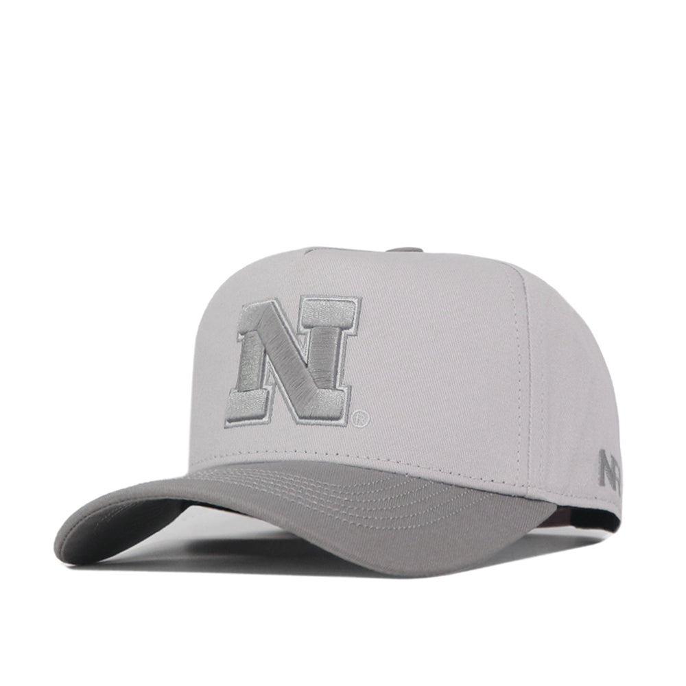 Introducing the Nebraska Graphite Hat by No Rivals—a light gray snapback cap featuring a raised, embroidered silver N logo on the front and a smaller N on the side. Made of fabric with a curved brim, this officially licensed hat combines classic style with team pride.